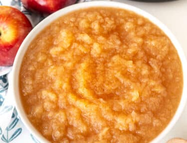Bowl of Instant Pot Applesauce in front of instant pot. Apples, cinnamon sticks and spoon on the side.