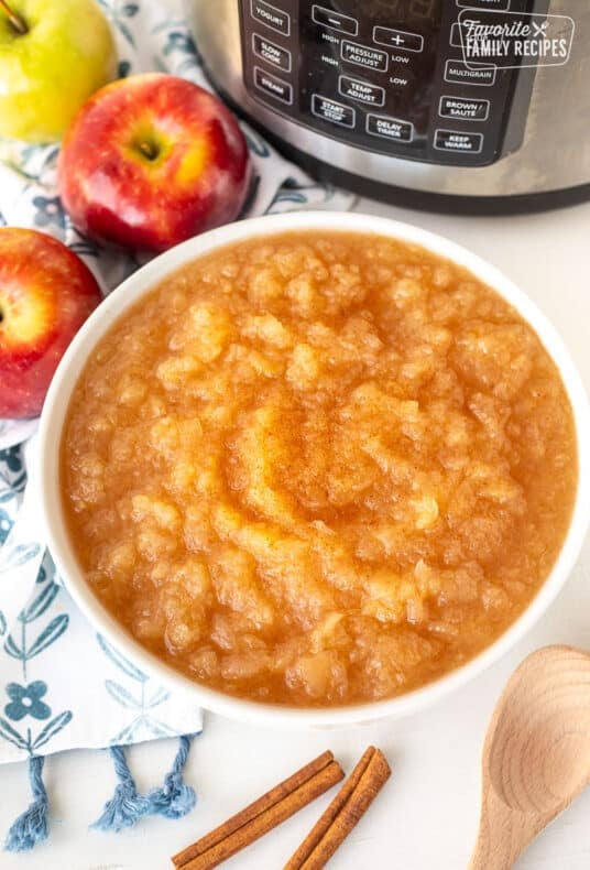 Bowl of Instant Pot Applesauce in front of instant pot. Apples, cinnamon sticks and spoon on the side.