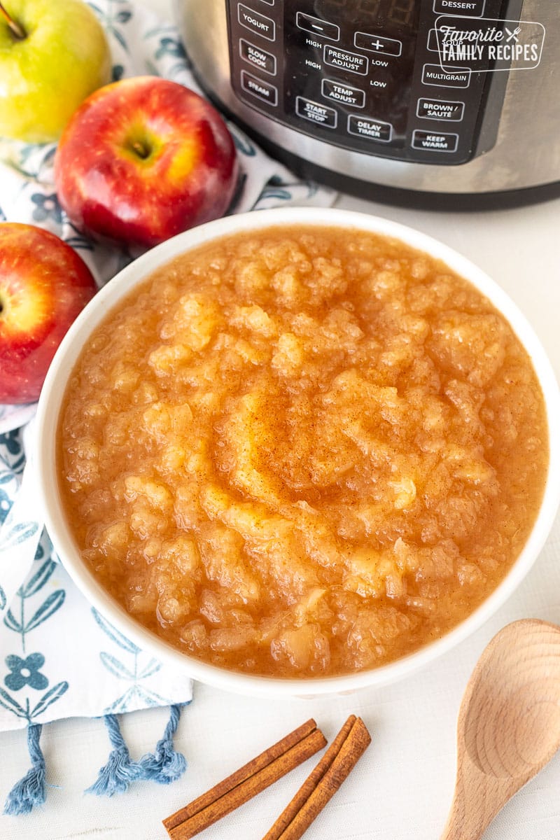 Bowl of Instant Pot Applesauce in front of instant pot. Apples, cinnamon sticks and spoon on the side.