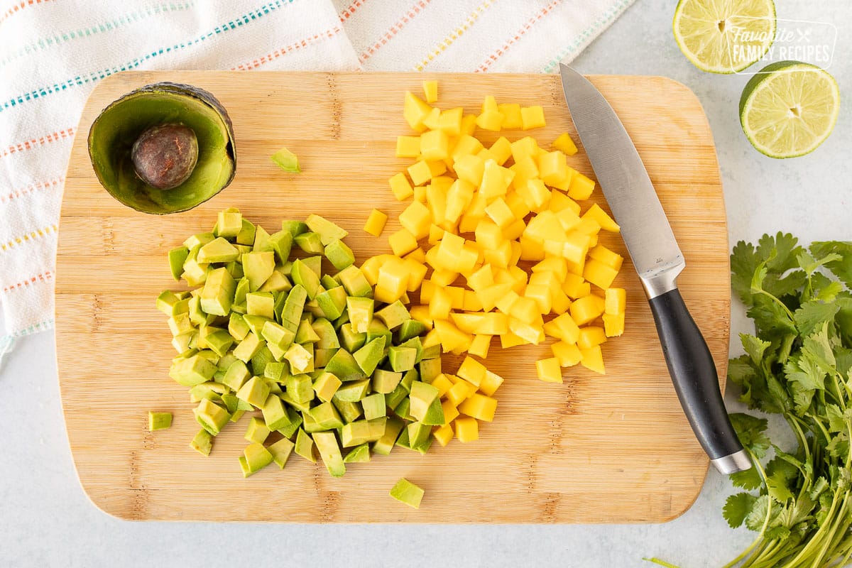 Cutting board of cut up avocados and mangos with a knife.