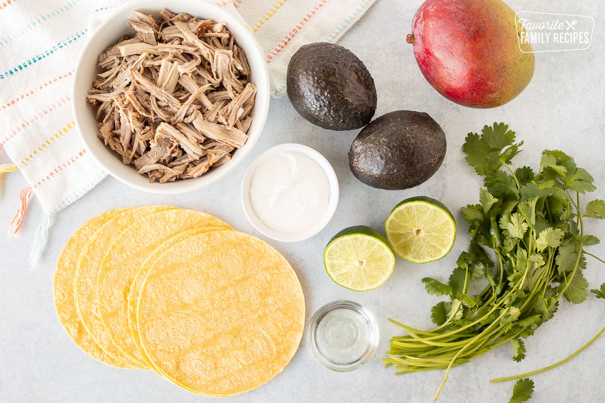 Ingredients to make Pulled Pork Tacos with Mango Salsa including tortillas, avocados, mango, cilantro, kalua pork, oil, lime and sour cream.
