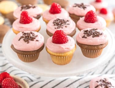 Vanilla and chocolate mini cupcakes frosted with raspberry frosting and topped with raspberries and chocolate sprinkles on a cake plate