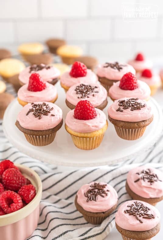 Vanilla and chocolate mini cupcakes frosted with raspberry frosting and topped with raspberries and chocolate sprinkles on a cake plate