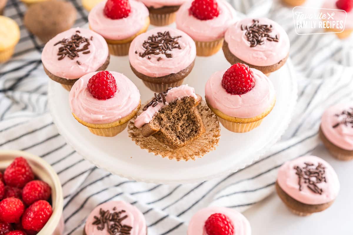 Vanilla and chocolate mini cupcakes frosted with raspberry frosting and topped with raspberries and chocolate sprinkles on a cake plate. One chocolate cupcake is unwrapped from the liner with a bite taken out of it.