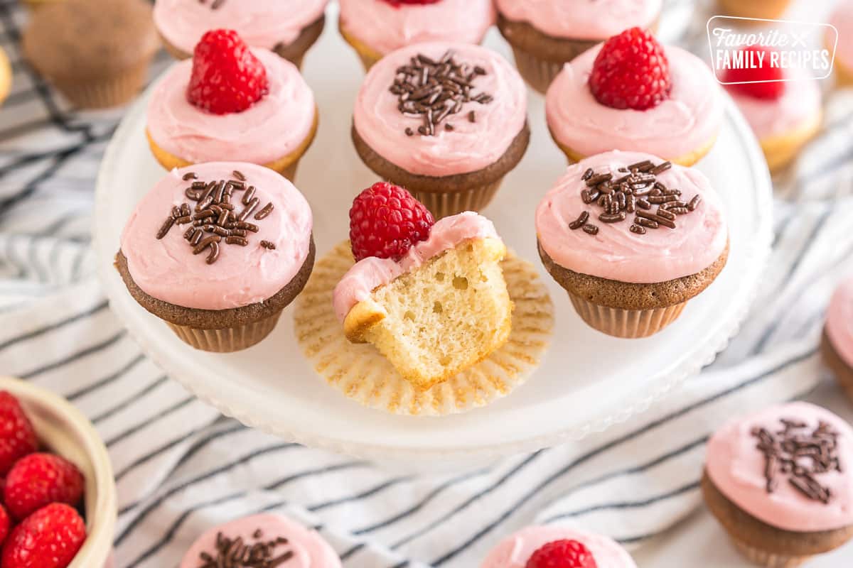 Vanilla and chocolate mini cupcakes frosted with raspberry frosting and topped with raspberries and chocolate sprinkles on a cake plate. One vanilla cupcake is unwrapped from the liner with a bite taken out of it.