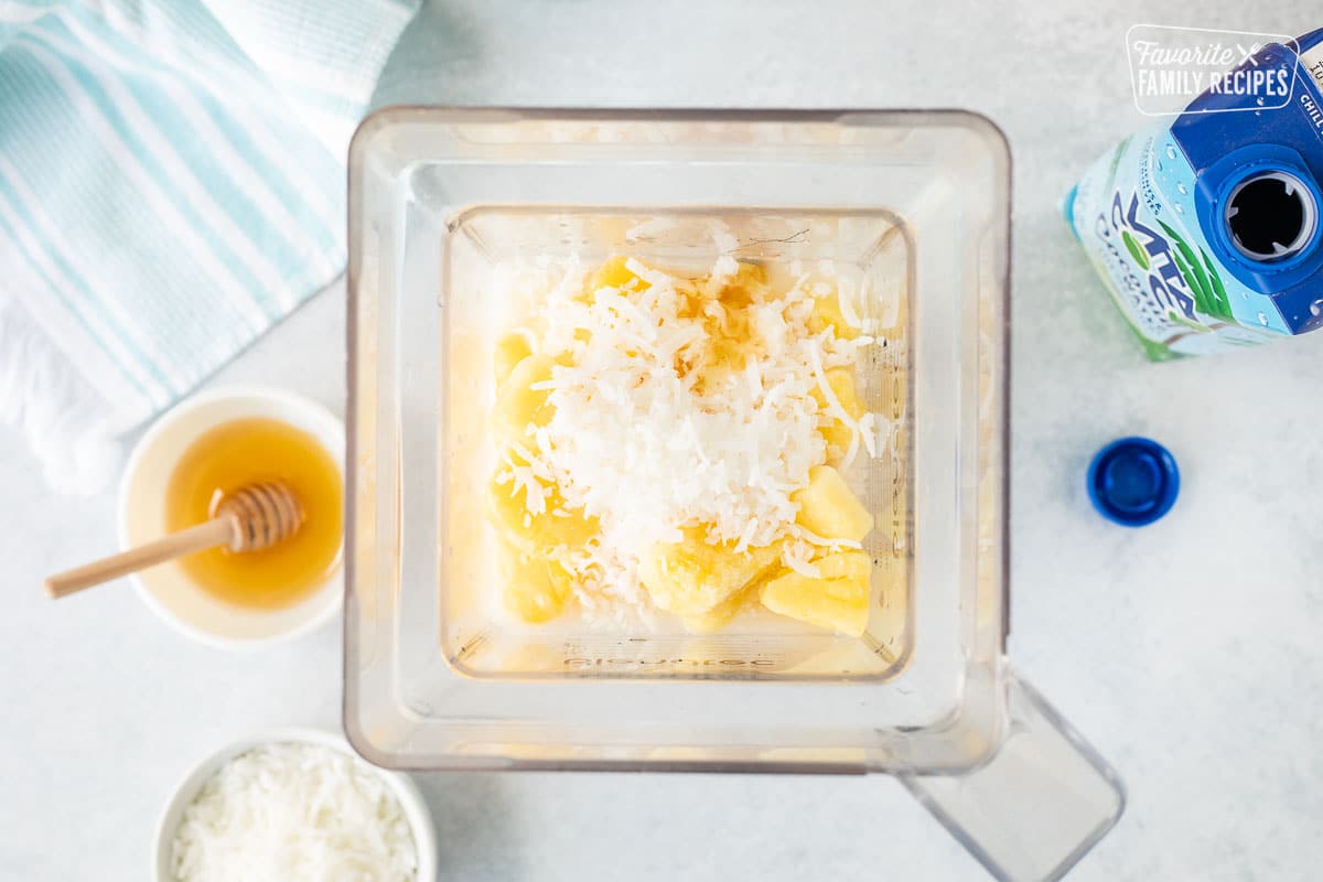 Blender with frozen pineapple, coconut, milk, honey and ice. Bowl of honey, coconut water carton and shredded coconut on the side.