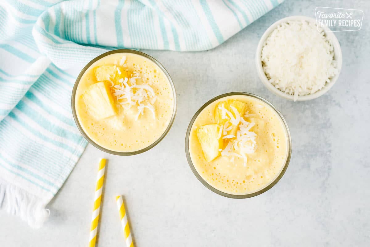 Two glasses of Pina Colada Smoothie with fresh sliced pineapple and shredded coconut on the side.