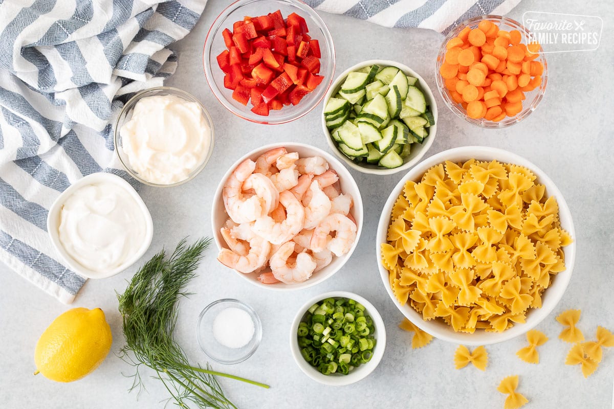 Ingredients to make Shrimp Pasta Salad including shrimp, mayonnaise, sour cream, red bell peppers, carrots, cucumbers, bow tie pasta, green onions, dill, salt and lemon.