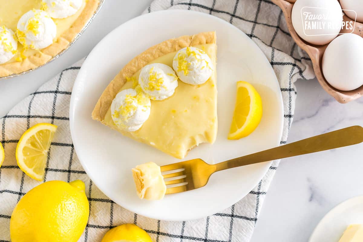 A slice of Sour Cream Lemon Pie on a plate topped with whipped cream and lemon zest with a fork taking a bite out
