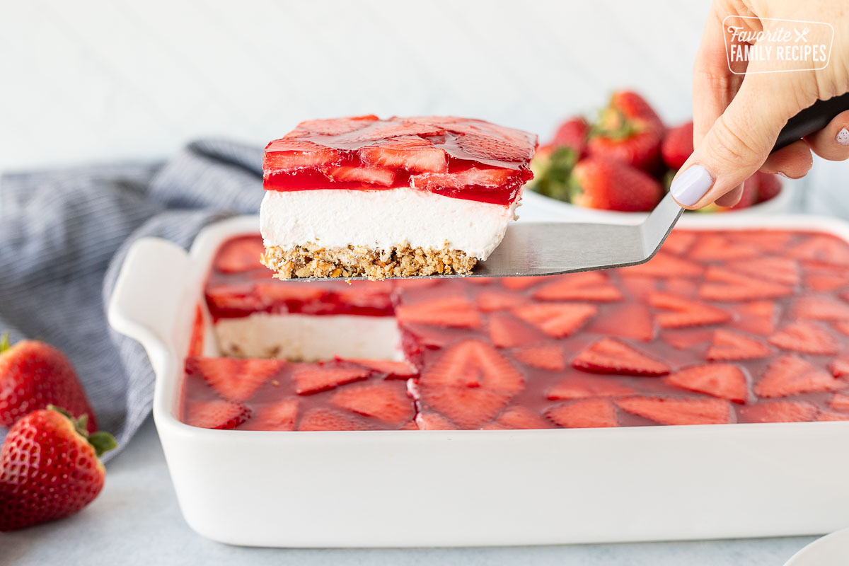 Spatula with a piece of Strawberry Pretzel Salad hovering above dish of Strawberry Pretzel Salad.