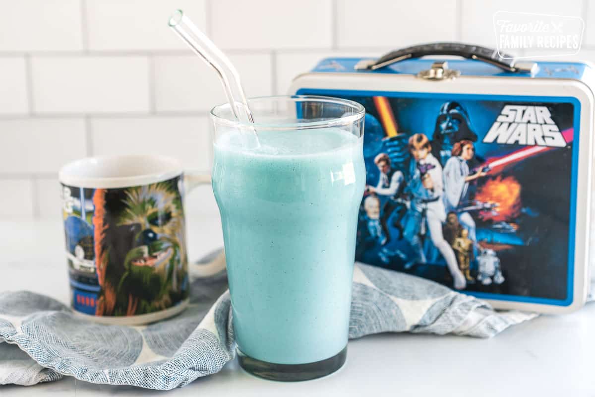 blue milk in a tall glass with a straw in front of a star wars lunchbox and a Chewy mug