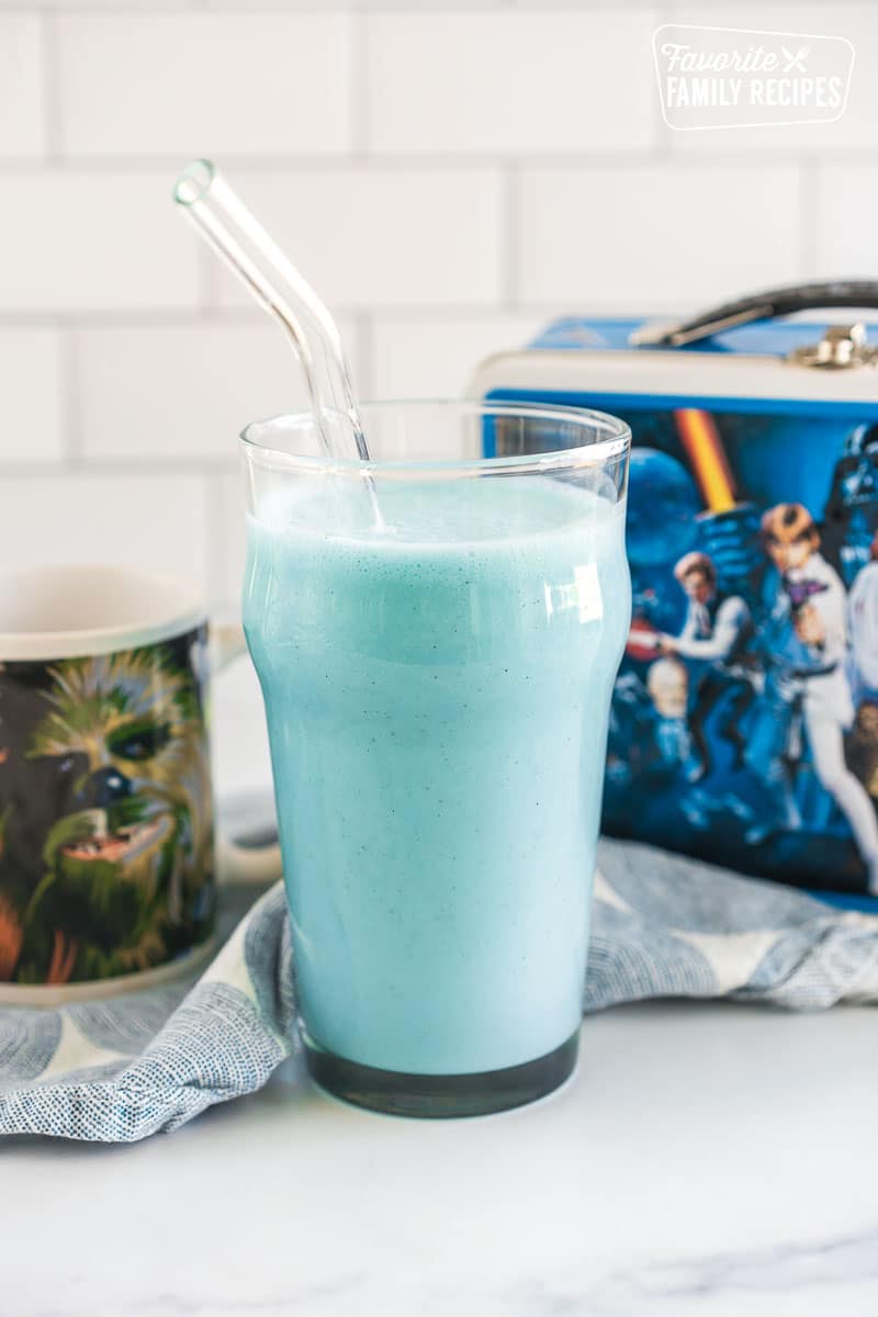 blue milk in a tall glass with a straw in front of a star wars lunchbox and a Chewy mug