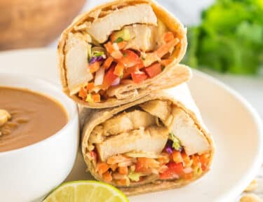 A thai chicken wrap cut in half on a plate next to a bowl of peanut sauce with a slice of lime