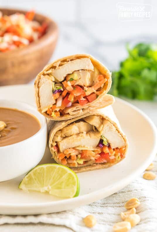 A thai chicken wrap cut in half on a plate next to a bowl of peanut sauce with a slice of lime