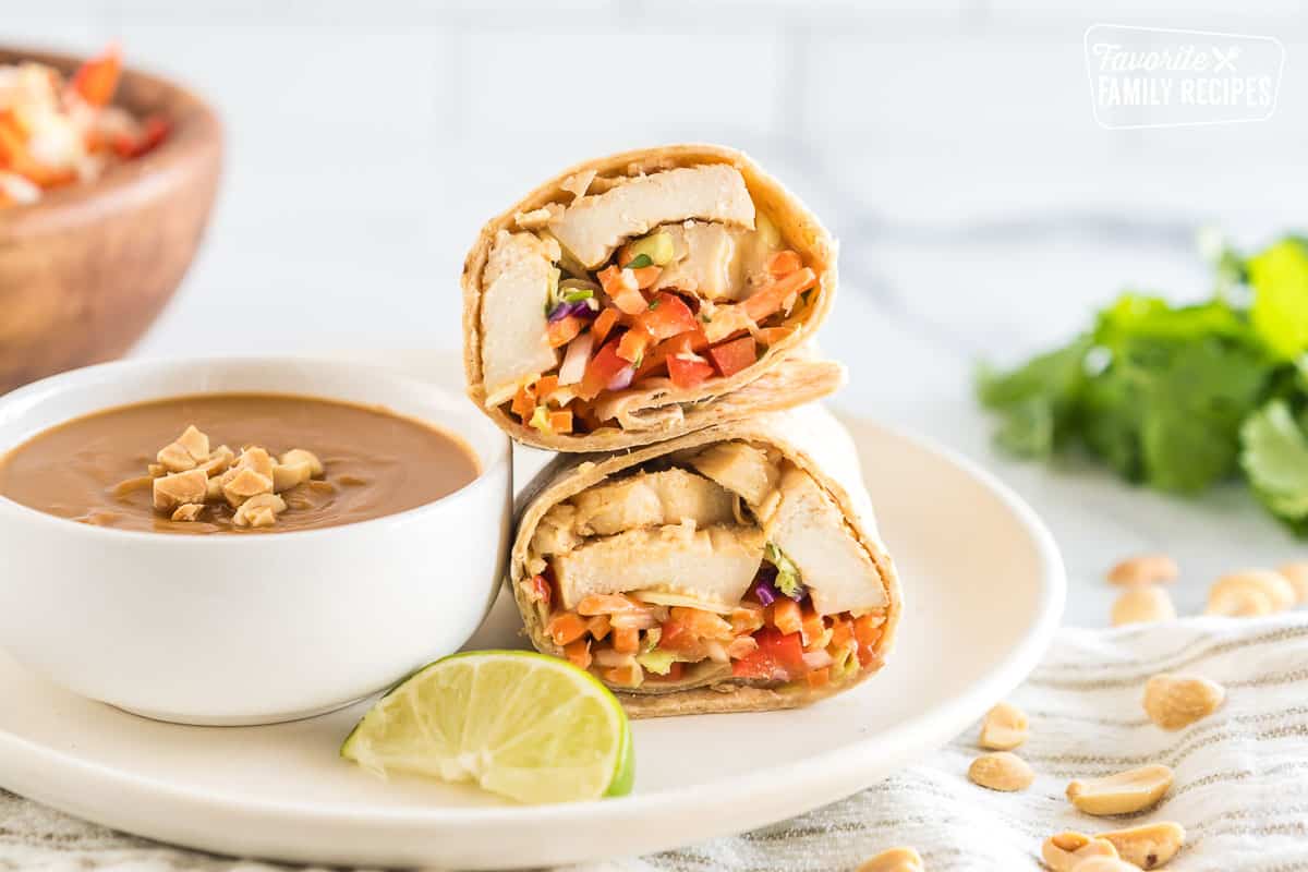 A Thai chicken wrap cut in half on a plate next to a bowl of peanut sauce with a slice of lime.