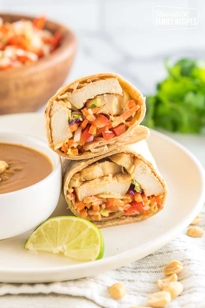A thai chicken wrap cut in half on a plate next to a bowl of peanut sauce with a slice of lime.