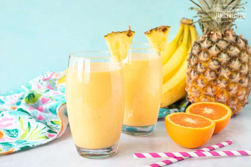 Two glasses of Tropical Banana Smoothie. Pineapple, bananas, oranges and straws on the side.