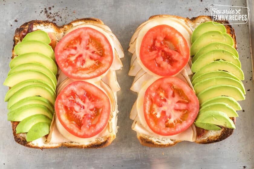 Two sub rolls sliced open and spread with cream cheese, meat, provolone cheese, tomato and avocado