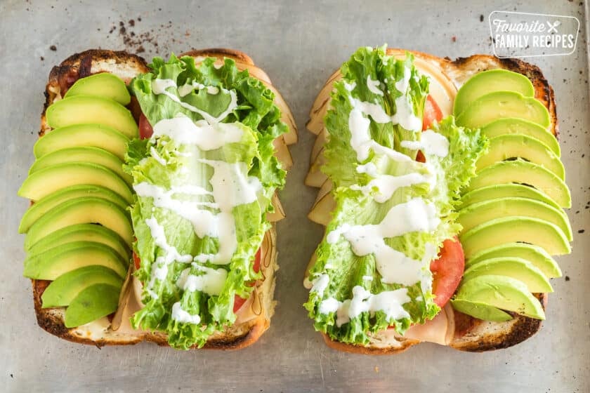 Two sub rolls sliced open and spread with cream cheese, meat, provolone cheese, tomato, avocado, lettuce, and ranch