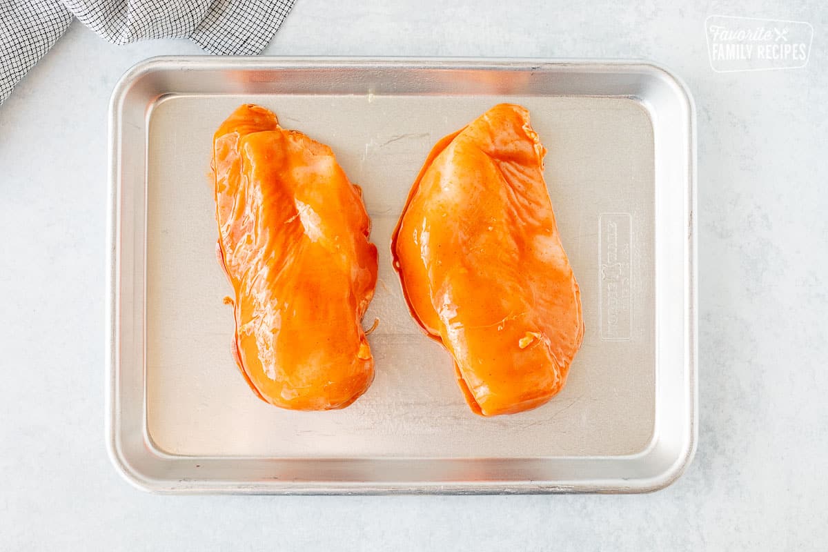 Two chicken breasts covered in Buffalo sauce on a baking sheet.