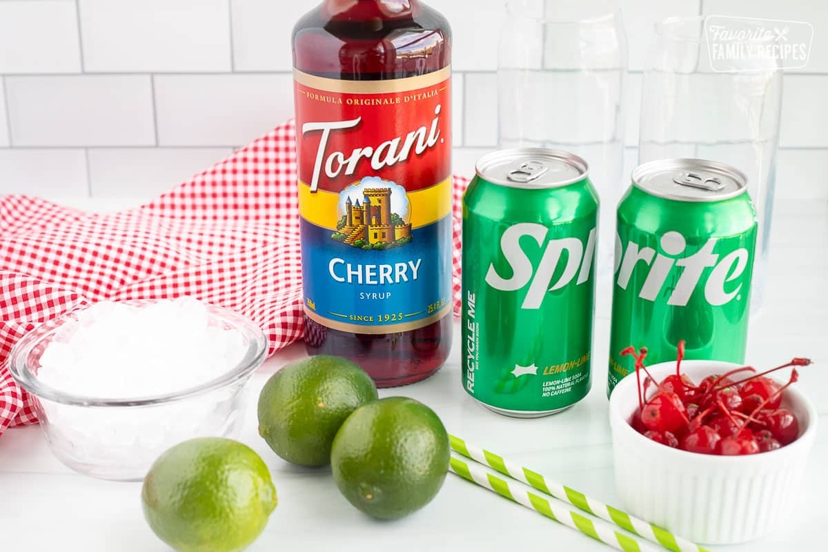 Ingredients to make Cherry Limeade including cherry syrup, Sprite, maraschino cherries, ice, lime, glasses and straws.