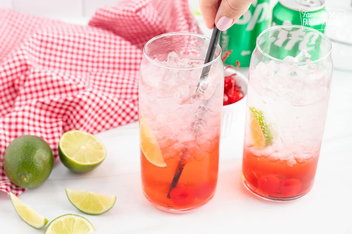 Two glasses of cherry limeade. Hand stirring one glass with a stir stick.