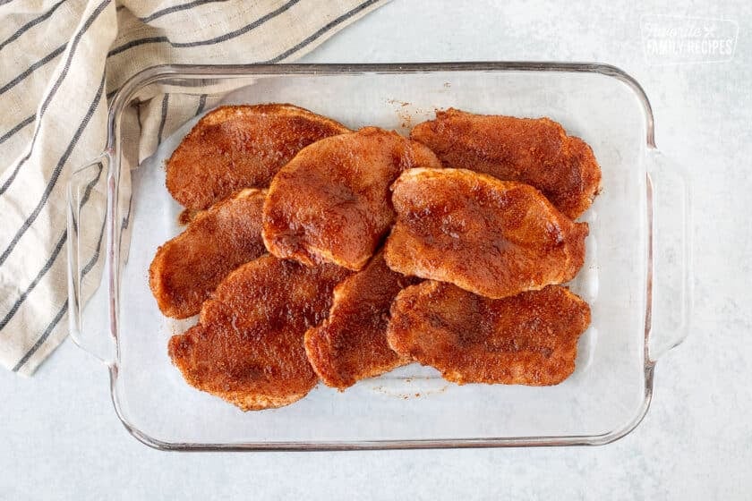 Glass dish of eight raw thin pork chops with seasoning rubbed all over each slice.
