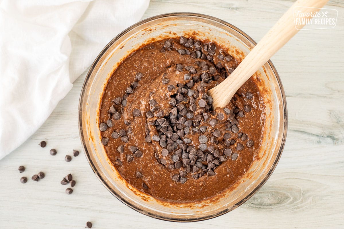 Wooden spoon mixing chocolate chip muffin batter in a glass bowl.