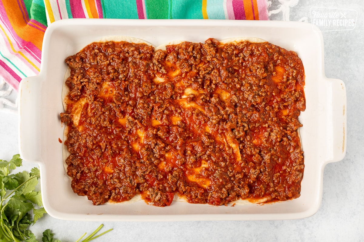 Glass baking dish with spread sauce on top of tortillas.