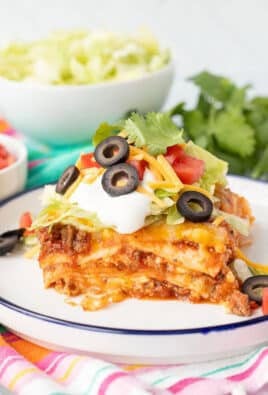 Slice of Mexican Lasagna on a plate with toppings including lettuce, cheese, tomatoes, sour cream, black olives and cilantro.