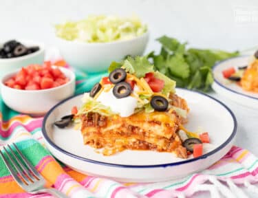 Slice of Mexican Lasagna on a plate with toppings including lettuce, cheese, tomatoes, sour cream, black olives and cilantro.
