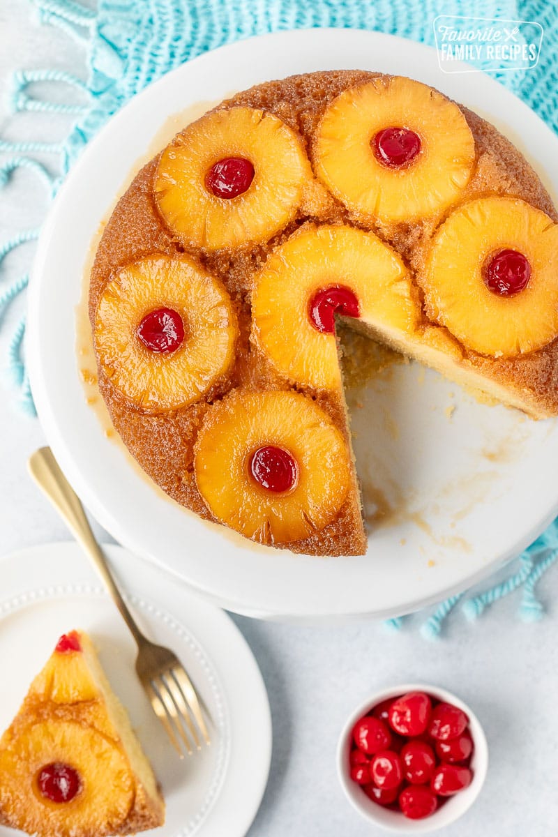 Pineapple Upside Down Cake on a cake stand next to a slice of cake and bowl of maraschino cherries.
