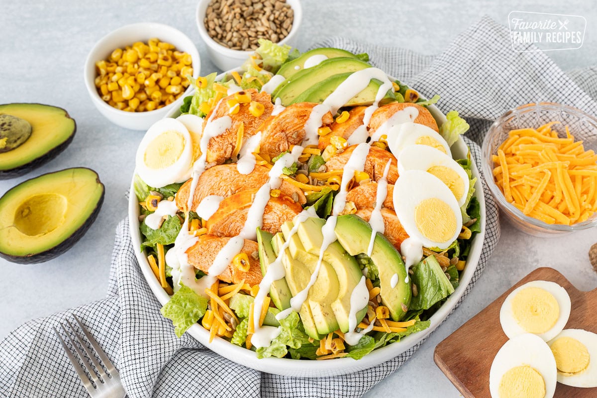 Bowl of Buffalo Chicken Salad topped with Ranch Dressing. Extra bowls of hard boiled eggs, avocado, cheese, grilled corn and sunflower seeds on the side.