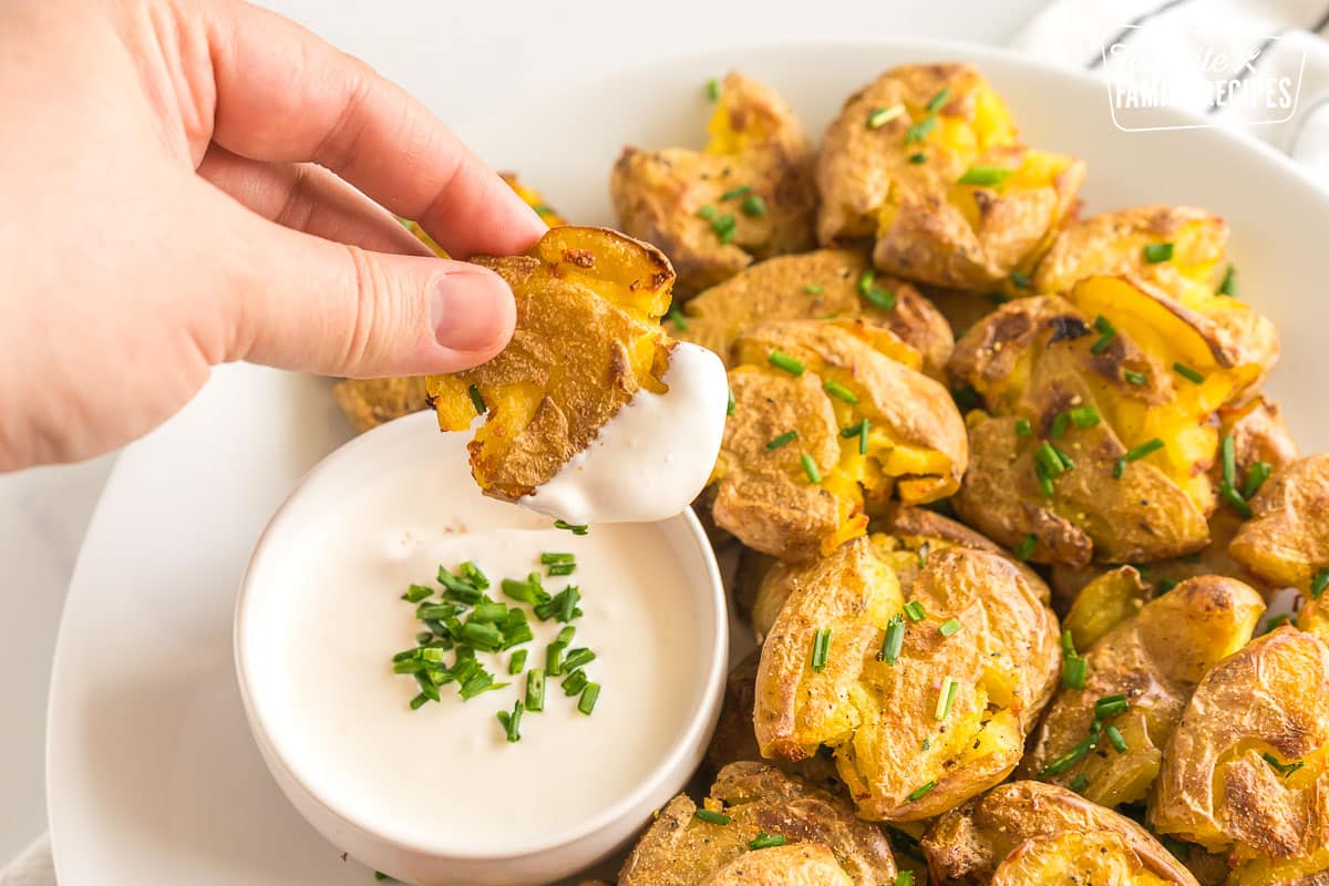 Smashed potatoes on a plate topped with chives. A hand is picking up one and dipping it in sour cream.