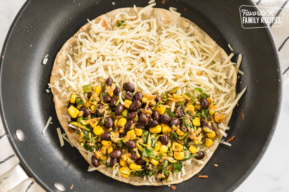 A tortilla in a skillet topped with cheese and a vegetable mixture.