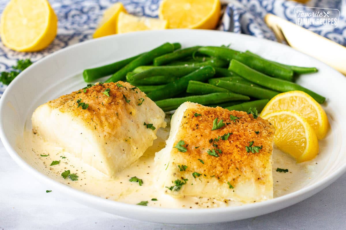 Plate with two pieces of Baked Cod in Cream Sauce next to green beans and lemon wedges.