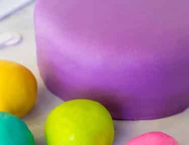 Covered fondant cake with purple fondant. Balls of pink, lime green, teal and yellow fondant balls around. Roller, cutter and smoother fondant tools on the side.