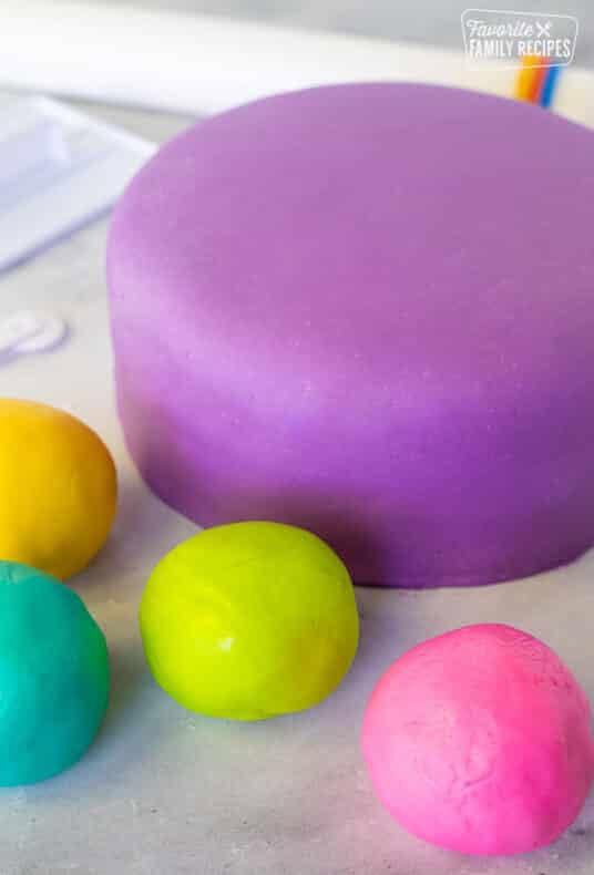 Covered fondant cake with purple fondant. Balls of pink, lime green, teal and yellow fondant balls around. Roller, cutter and smoother fondant tools on the side.