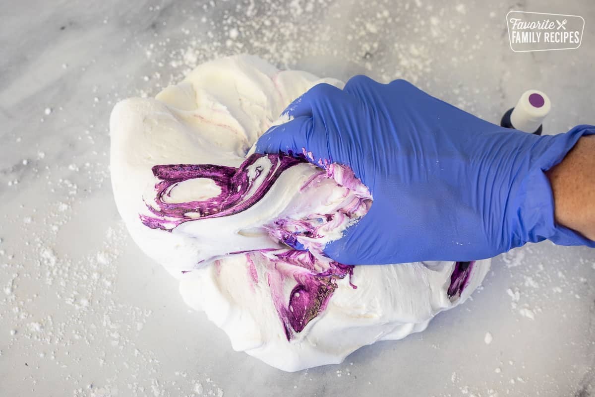 Mixing purple gel coloring into a ball of fondant with a gloved hand.
