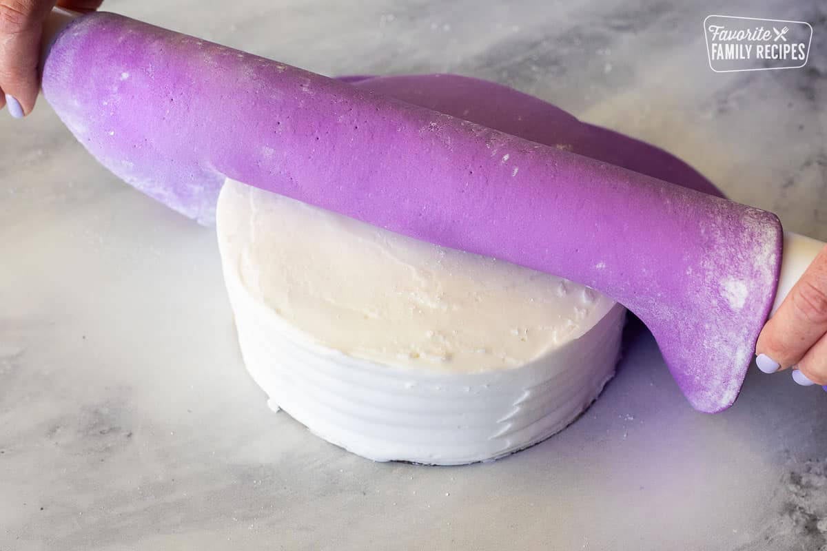Rolling a sheet of purple fondant on top of a white frosted cake.
