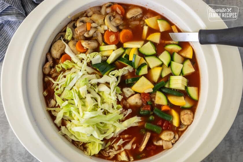 Crock pot with tomato broth, sliced zucchini, yellow squash, carrots, cabbage, mushrooms and spoon.