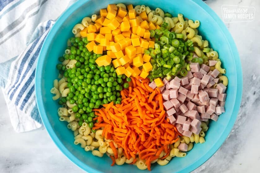 Bowl of cooked pasta with green peas, cubed cheese, sliced green onions, cubed ham and shredded carrot.