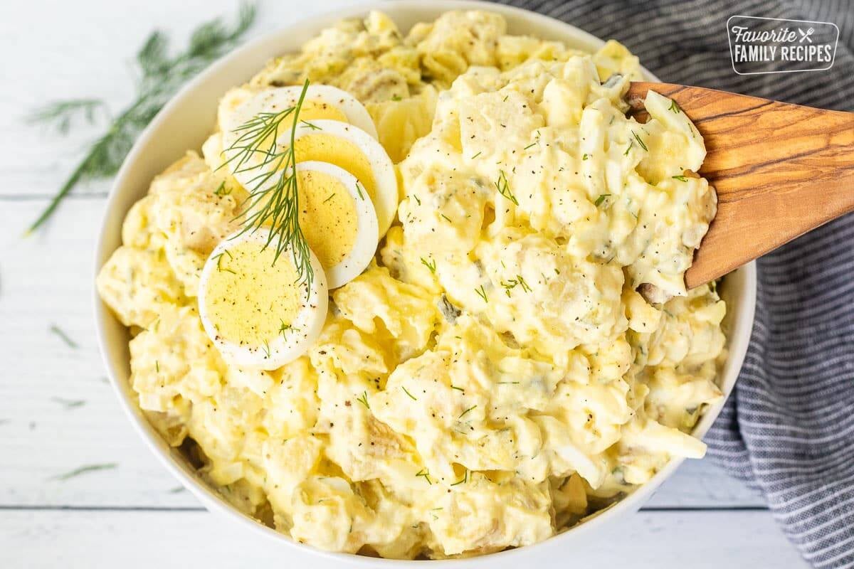 Wooden spoon scooping from a bowl of Homemade Potato Salad with sliced hard boiled eggs on top and fresh dill.