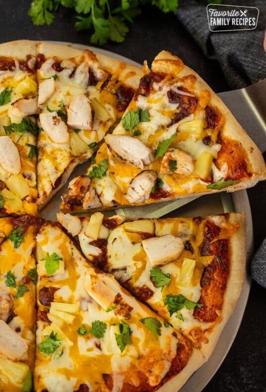A slice of barbecue chicken with chicken, cilantro, and cheese being served from the full pizza pie