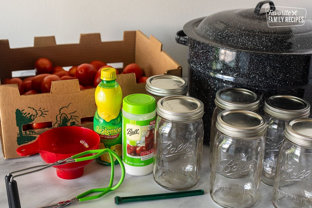 Ingredients to can whole tomatoes including a box of Roma tomatoes, lemon juice, citric acid, canning funnel, jar lifter, lid lifter, canning jars and a canning pot.