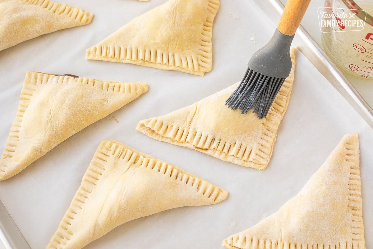 Brushing egg wash on tops of unbaked apple turnovers on a baking sheet covered with parchment paper.