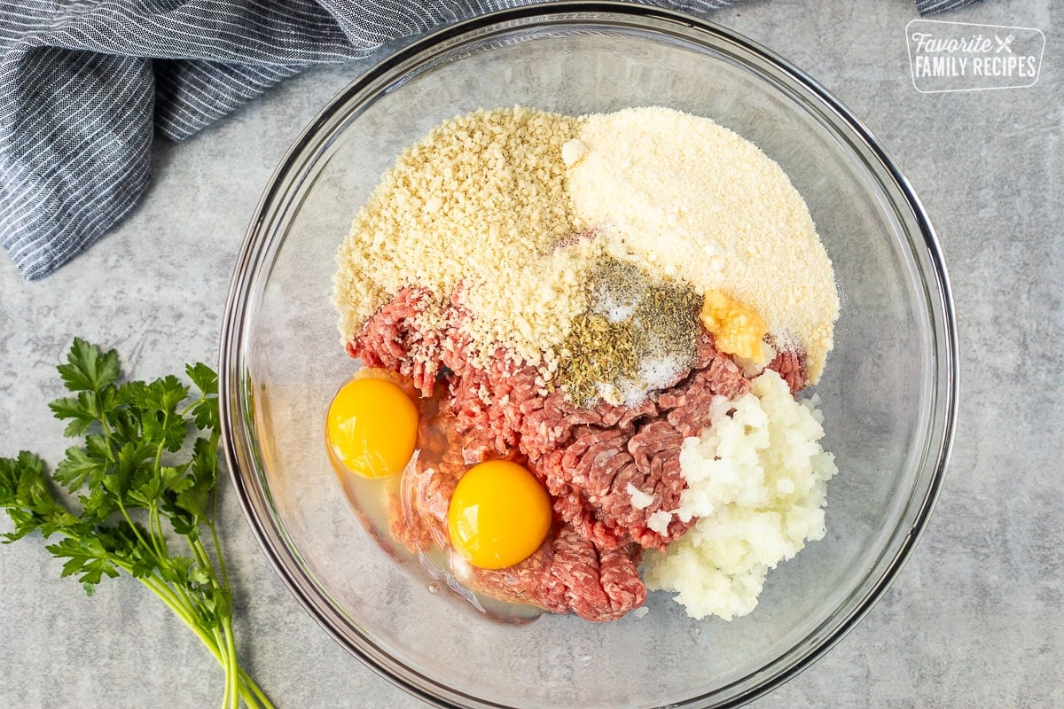 Glass mixing bowl with ground beef, cracked eggs, onion, garlic, Parmesan cheese, seasonings, and panko crumbs.