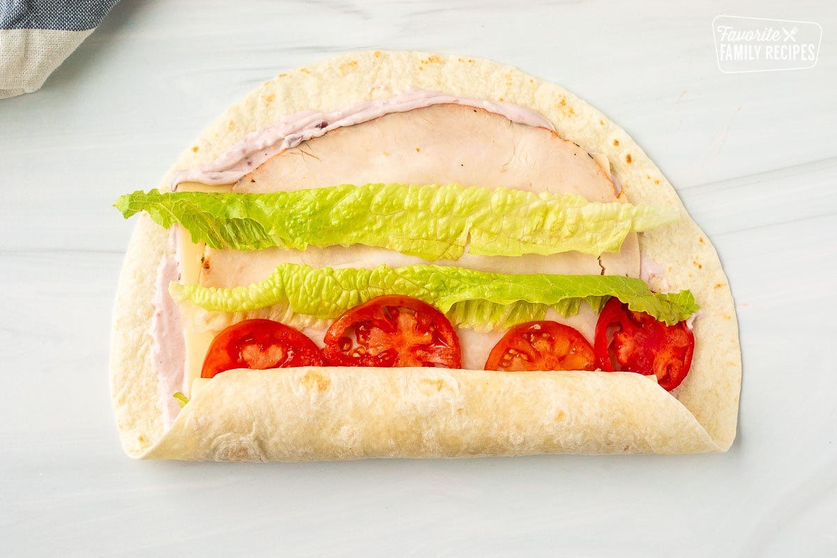 Halfway rolled up tortilla for a turkey roll up with cranberry cream cheese, Swiss cheese, turkey slices, lettuce and tomatoes.