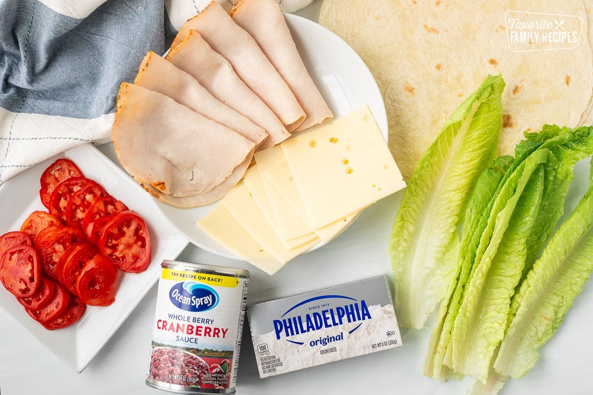 Ingredients to make turkey roll ups including lettuce, tortillas, cream cheese, sliced tomatoes, cranberry sauce, turkey slices, and Swiss cheese.