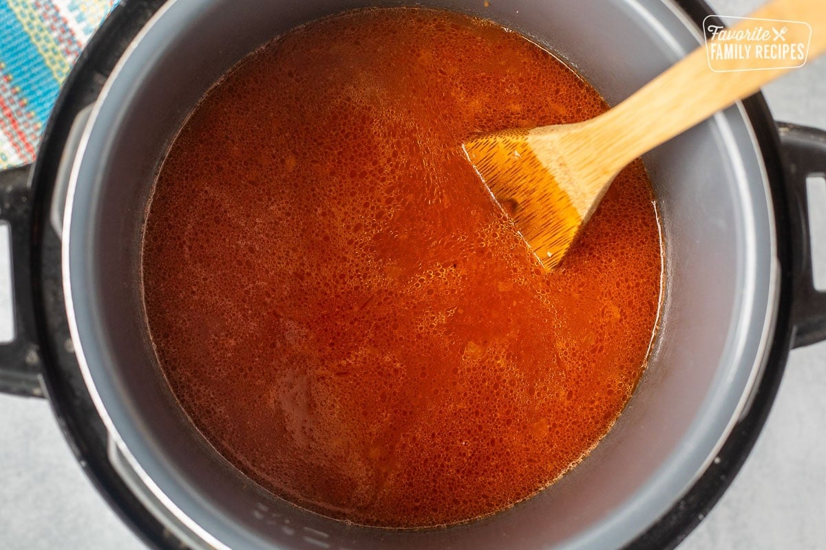 Instant pot with tomato sauce and chicken broth combined with rice and a wooden spoon stirring.